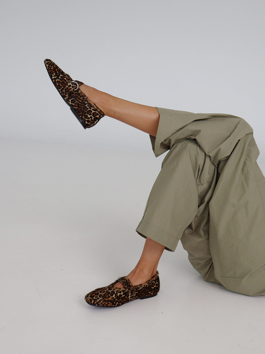 A beige, black, and brown animal patterned flat shoe with an adjustable Mary-Jane strap on a model with crossed legs.