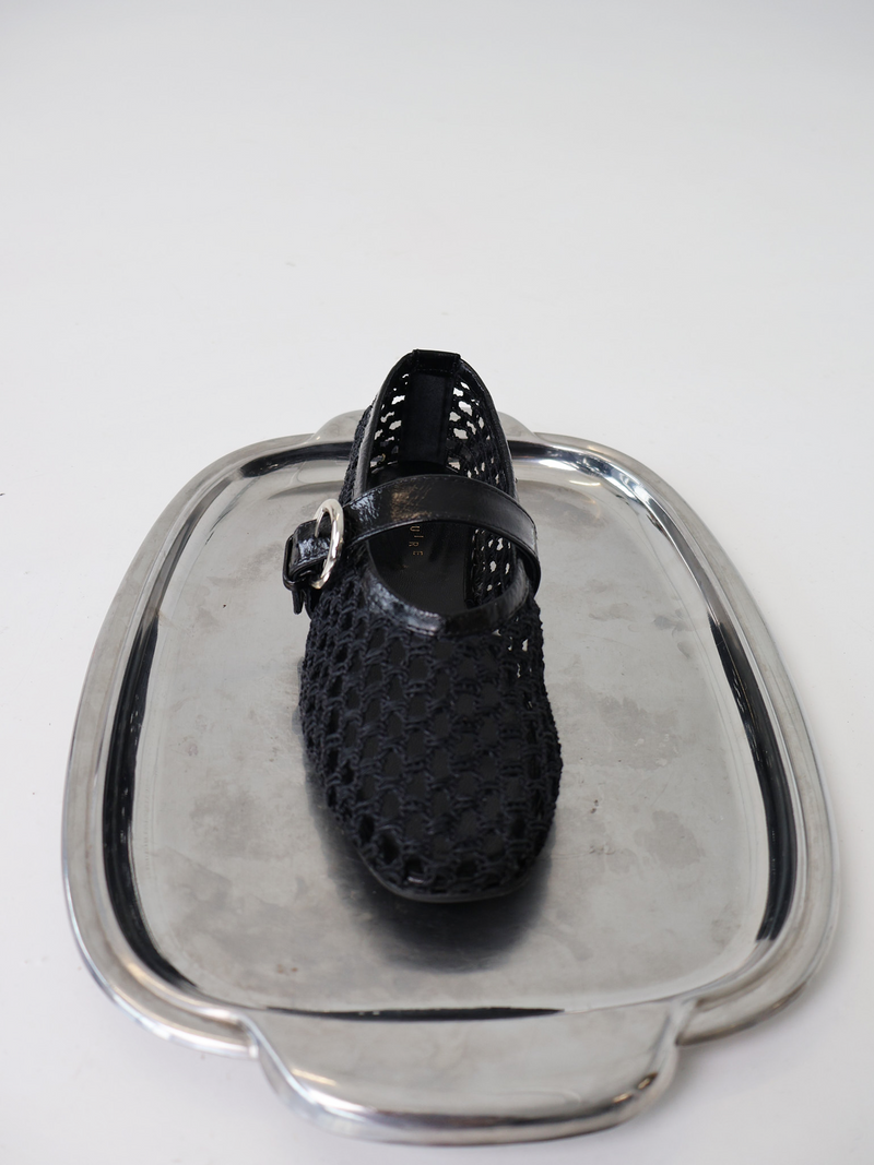 A black woven ballerina flat with an adjustable Mary-Jane strap, displayed on a silver metallic plater