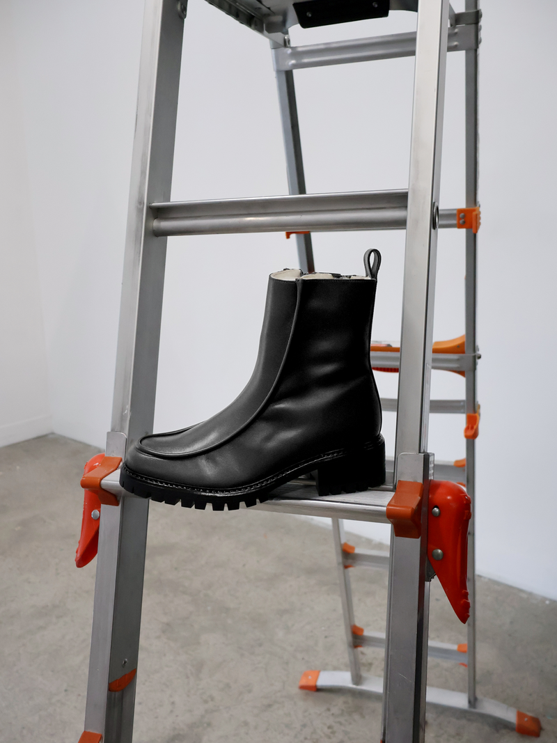 Black lined winter boot with rounded toe, 3d piping detailing and a back tab sitting on a ladder.