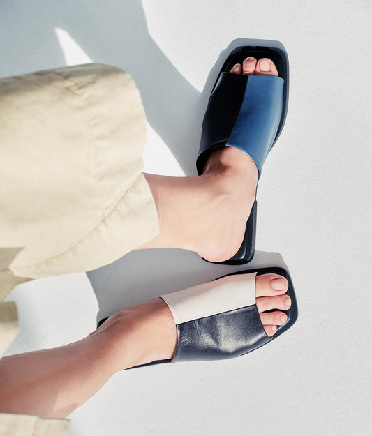 Sandal with for colours on model wearing a beige skirt. The shoes are Black and blue on one side and white and black on the other. Mismatch pair.