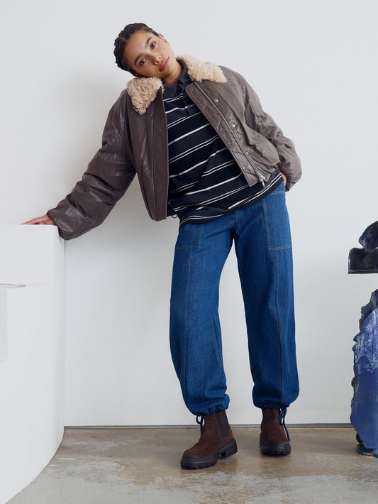 Woman wearing brown suede chelsea boots, standing against white wall and wearing some balloon pant jeans with a striped shirt and pleather brown jacket.