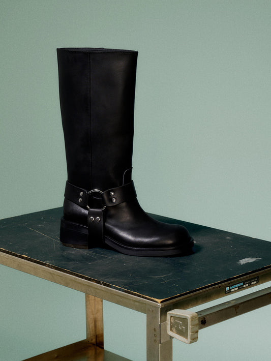A black leather boot with a harness detail and block heel, on a green background.