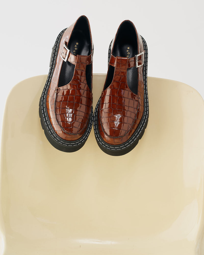 Brown crocodile-patterned shoes with black soles on a beige chair.