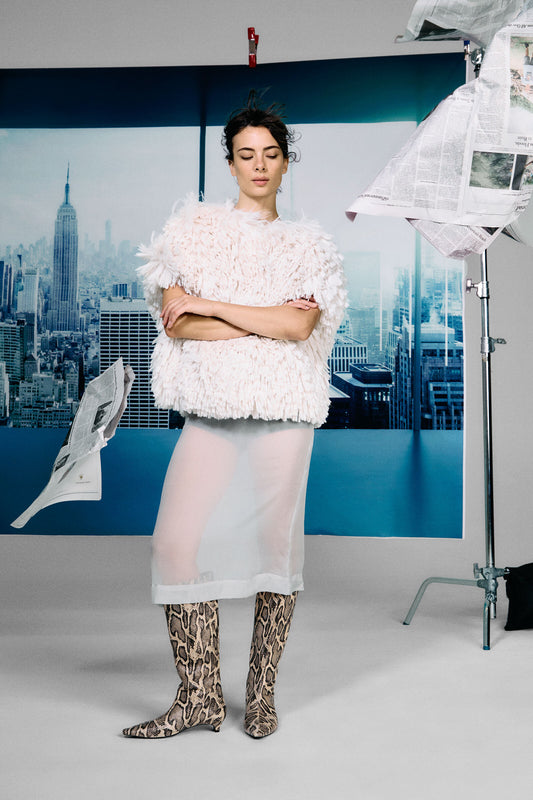 Model wearing a textured white top and sheer skirt with snake print boots in a studio setting.