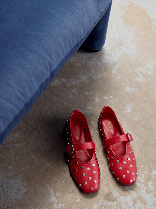 Red ballet flat with Mary-Jane strap and silver studs next to a blue sofa.