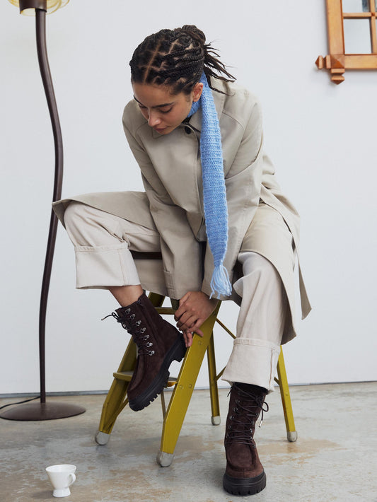 A brown suede chunky combat boot with lace-up closure and metal hardware worn with a nude outfit.