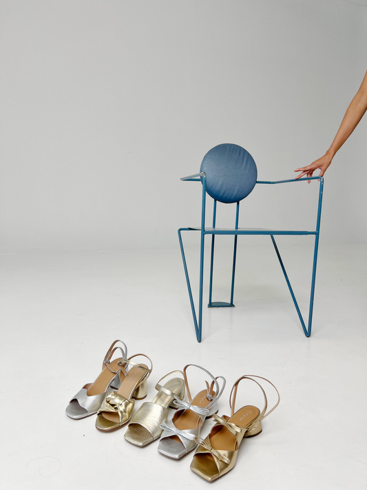 Gold high-heeled sandals on a white surface with a blue chair in the background.