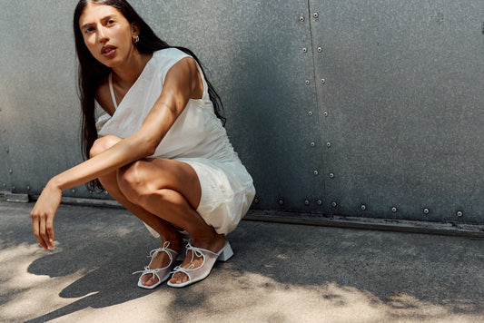 Woman wearing a white dress with white heeled sandals crouching at a street corner