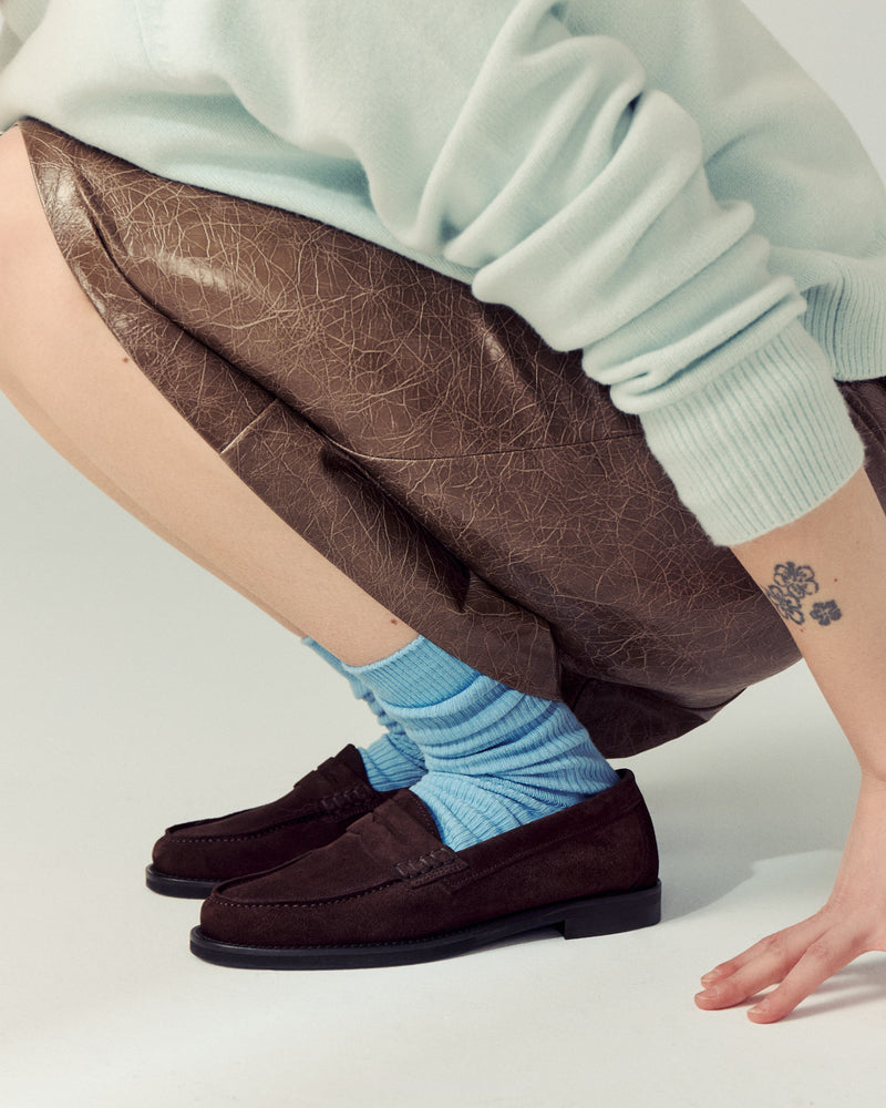 woman crouching down wearing brown skirt, mint green sweater, blue socks and brown suede loafers