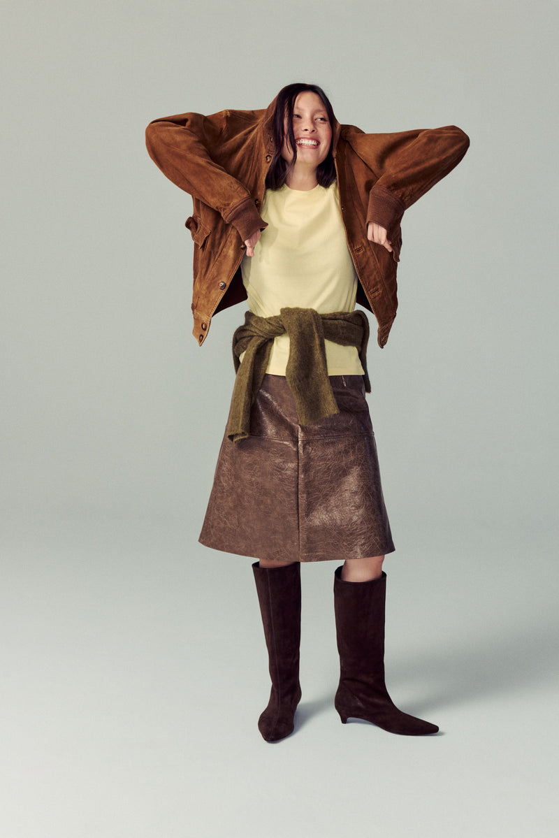 girl standing in white studio wearing brown suede heeled boots, brown skirt, yellow top with green sweater at her waist, smiling and playing around with jacket