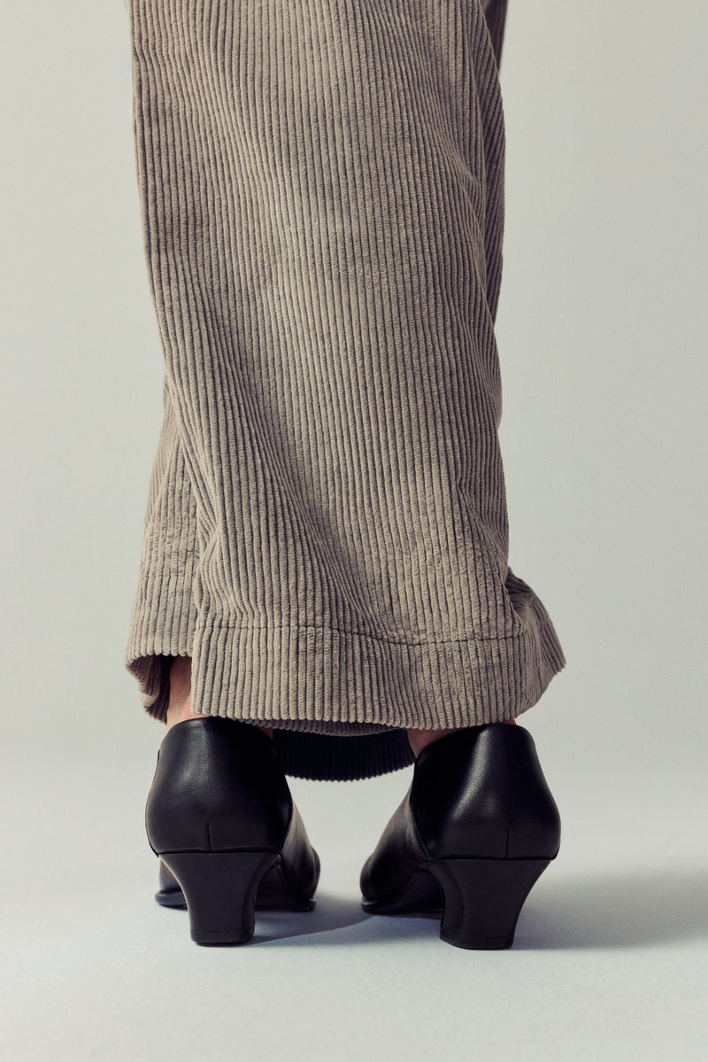 backshot of woman standing crossing her legs, wearing corduroy taupe pants and black heels