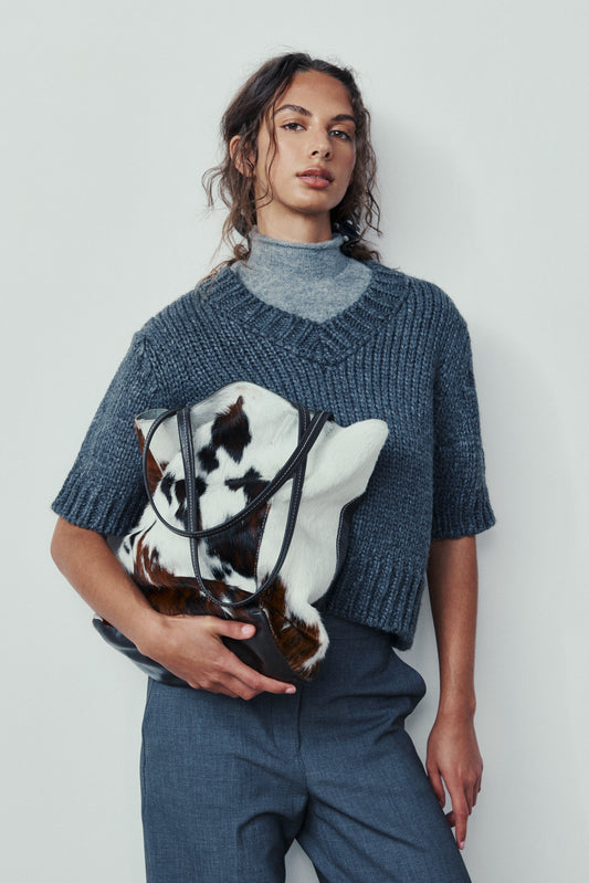 Person wearing a blue knitted sweater holding a cow-patterned bag against a plain background