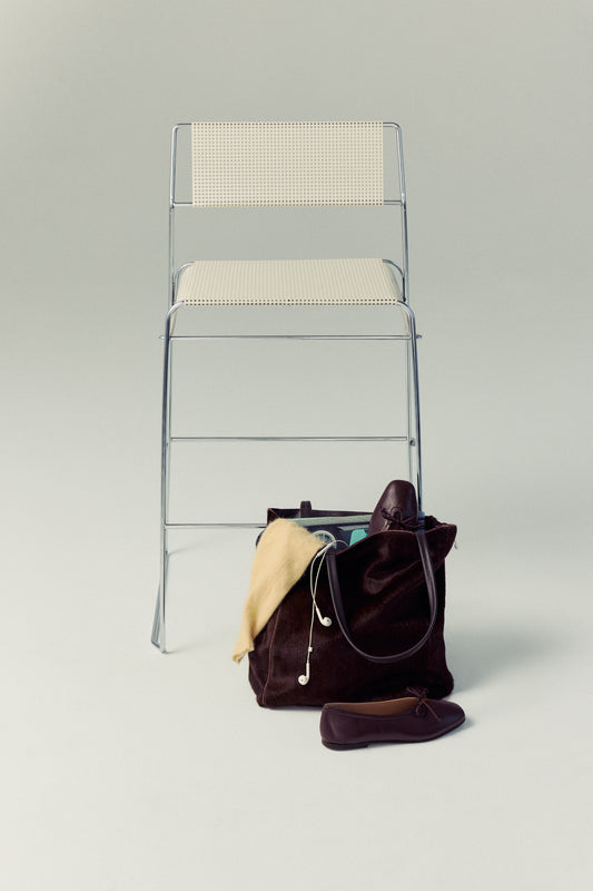 Clear chair with a beige cushion next to a brown Carpi handbag on a light gray background