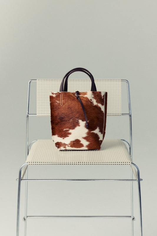 Brown and white cowhide Carpi leather bag on a clear chair with a gray background