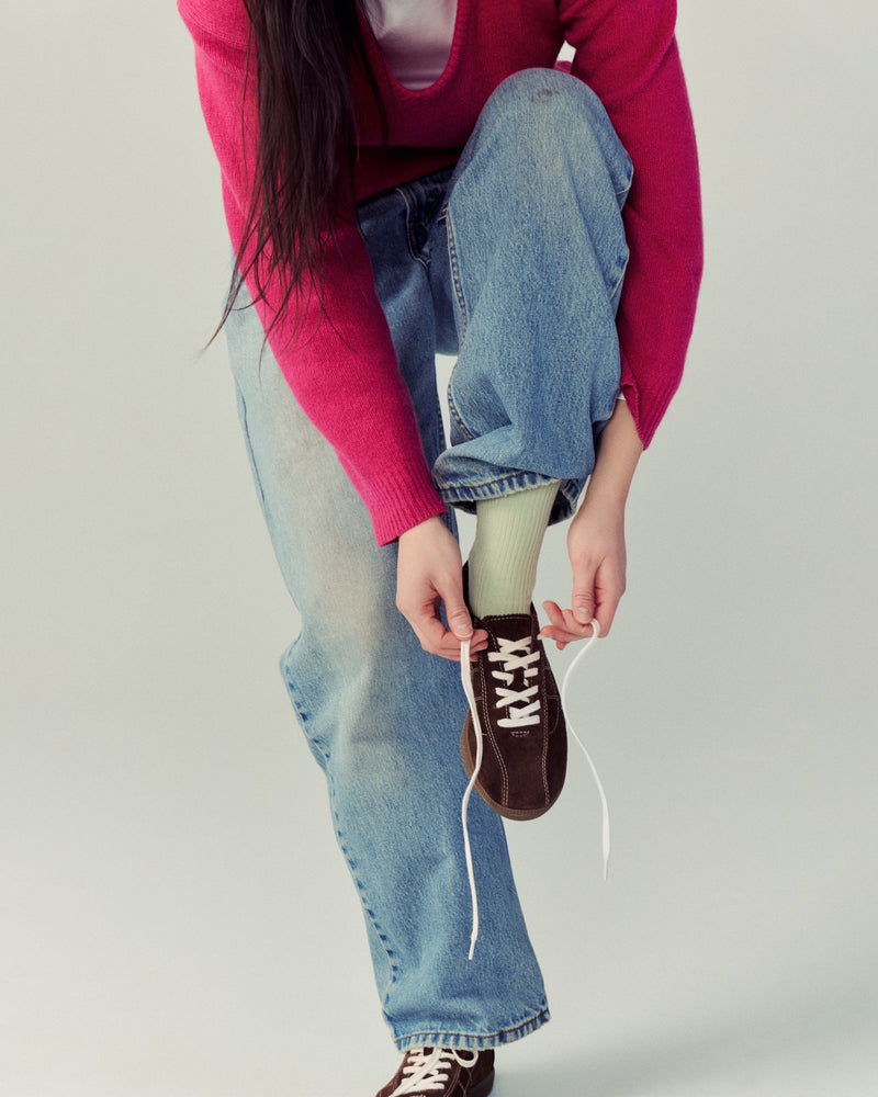 Person wearing a pink sweater and blue jeans, holding a Simone brown sneaker against a plain background