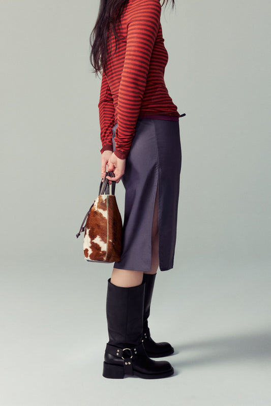 Woman standing in black leasther biker boots, holding a cow printed mini bag, with long brown hair and a full outfit from paloma wool (striped red shirt and purple skirt)