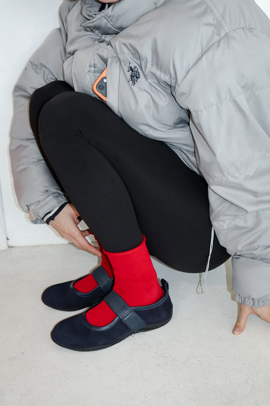 Woman crouching on white floor, wearing black leggings, gray puffer, and navy flats.