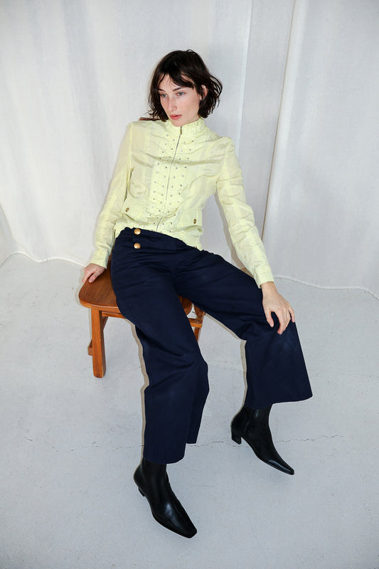 Woman sitting on a stool wearing a light green sweater and dark blue pants against a white background
