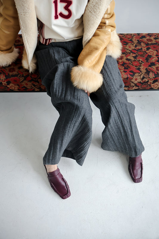 Person wearing a coat with fur trim, white shirt with red number 13, gray ribbed pants, and bordeaux shoes on a white floor.