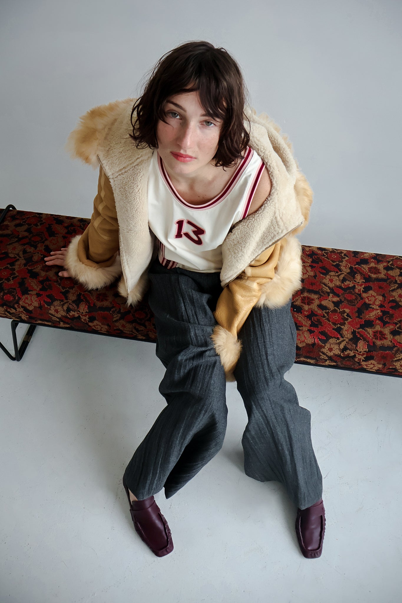 Person wearing a vintage outfit with a fur-trimmed coat, white shirt with red number 13, and gray pants, sitting on a patterned bench with Bordeaux shoes.