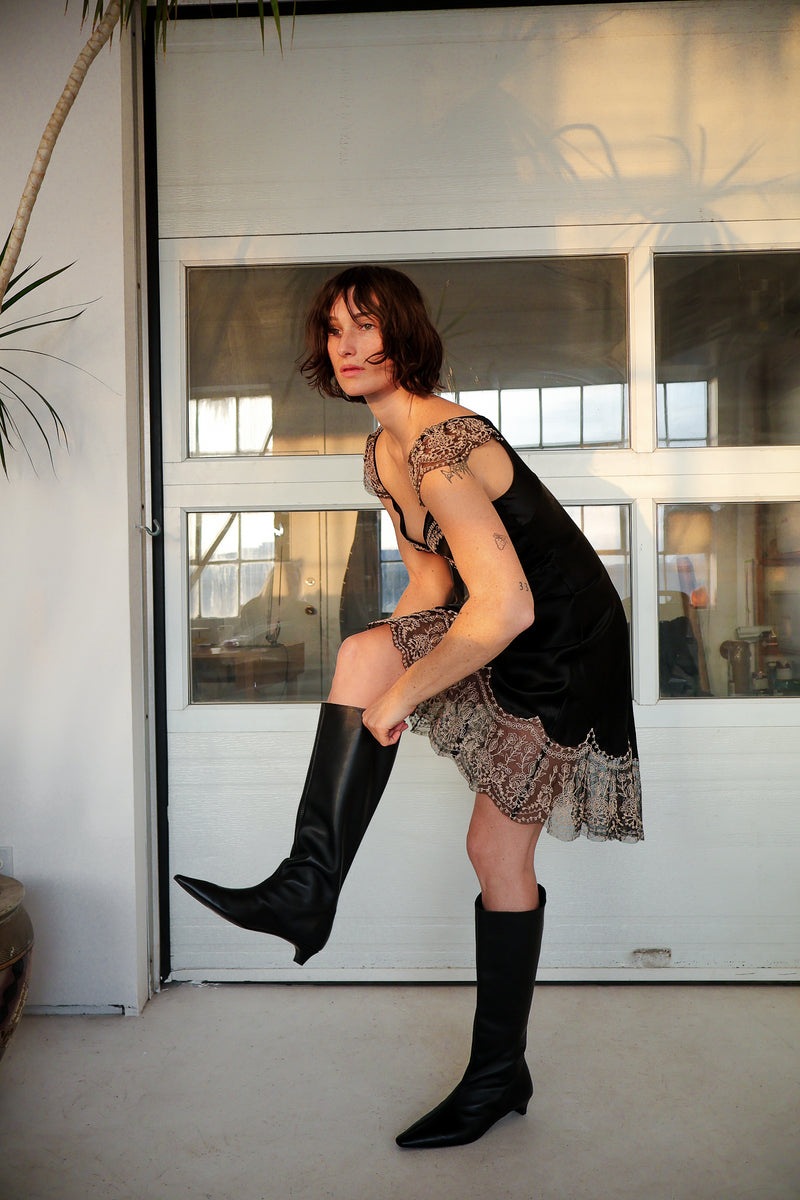 Woman in black lace dress and knee-high boots posing indoors.