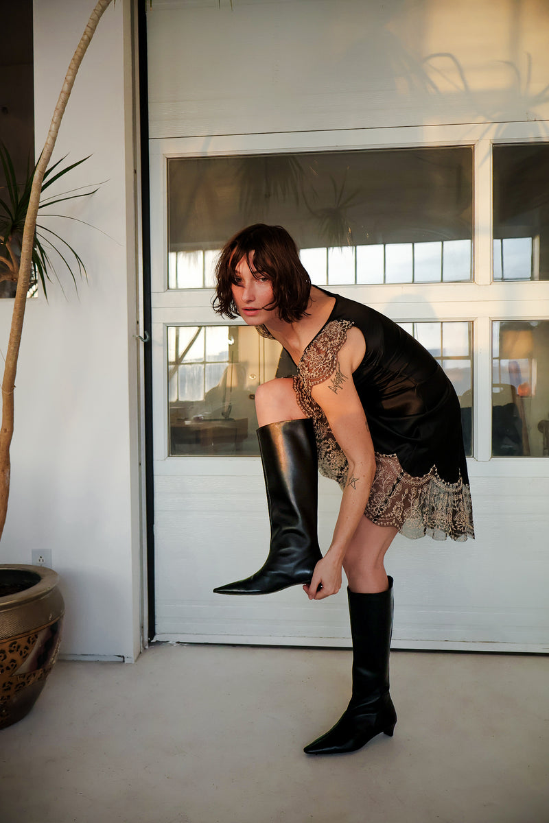 Woman wearing black knee-high boots and a black top with lace details in a modern indoor setting.