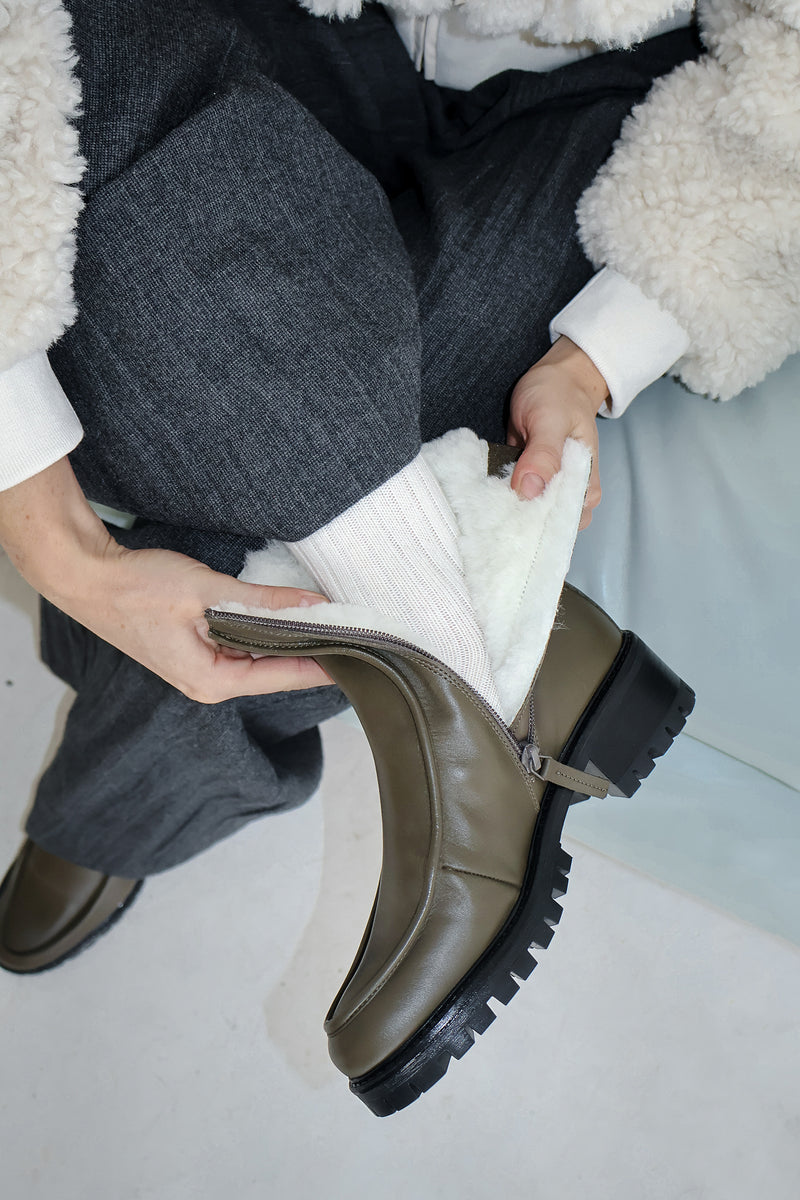 Woman is wearing a shearling lined winter boot in an olive green leather. She's opening the boot so we see the softness of the white sheepskin shearling. 