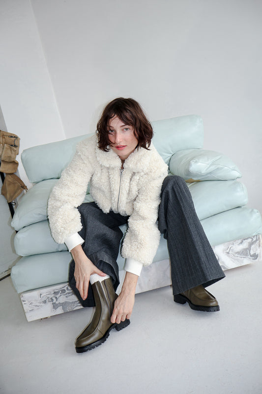 Woman sitting on a couch wearing a shearling lined winter boot in an olive green leather. Boot is unzip and we can see the soft shearling lining. 
