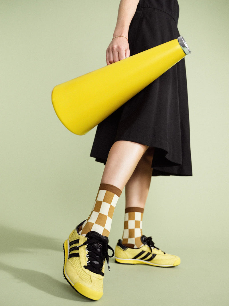 A person wearing a black skirt and yellow checkered socks, with yellow sports shoes featuring black accents, standing on one leg holding a yellow megaphone.