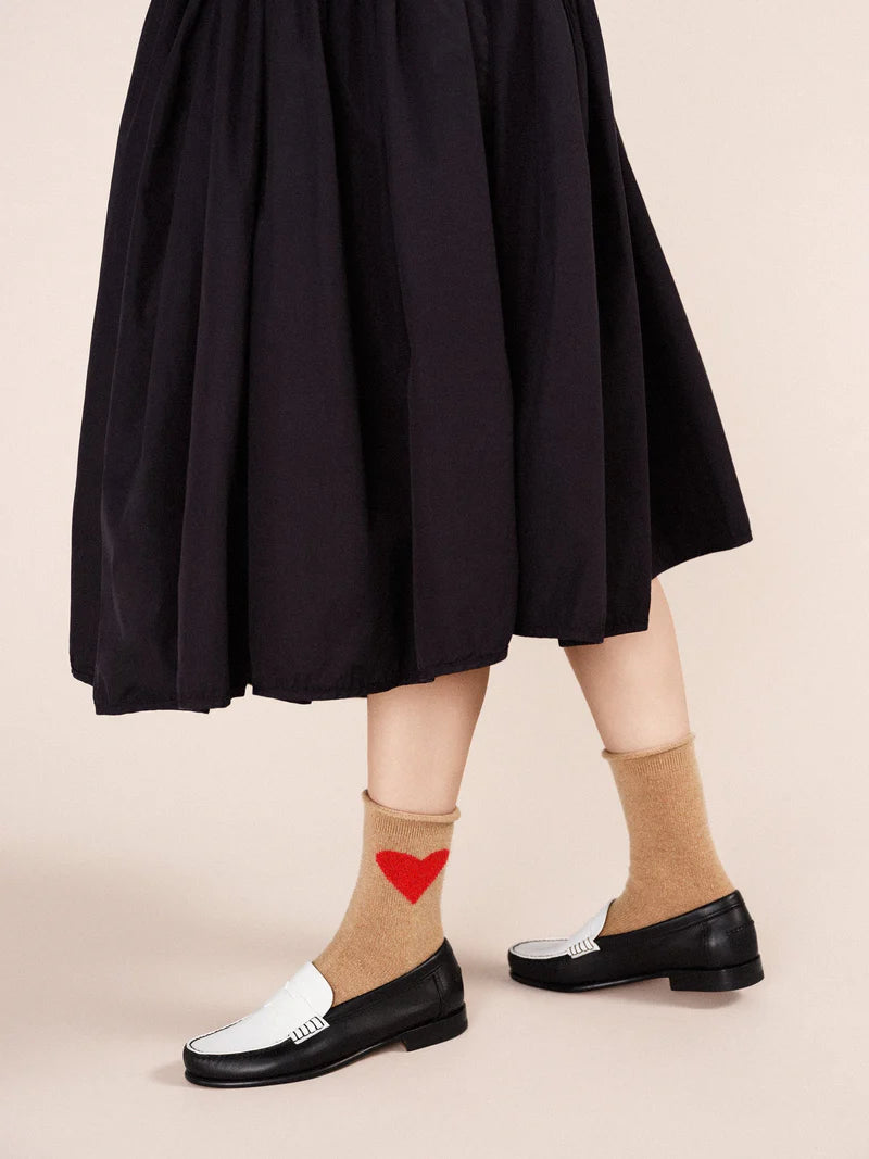A person wearing beige cashmere socks with a red heart motif, with black and white shoes and a dark skirt.
