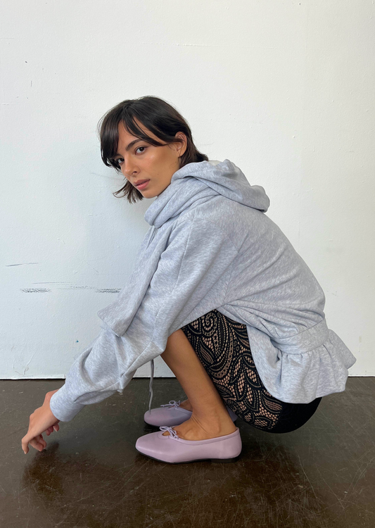 Woman wearing a gray hoodie and black pants with lace details, sitting on a dark surface against a white wall.
